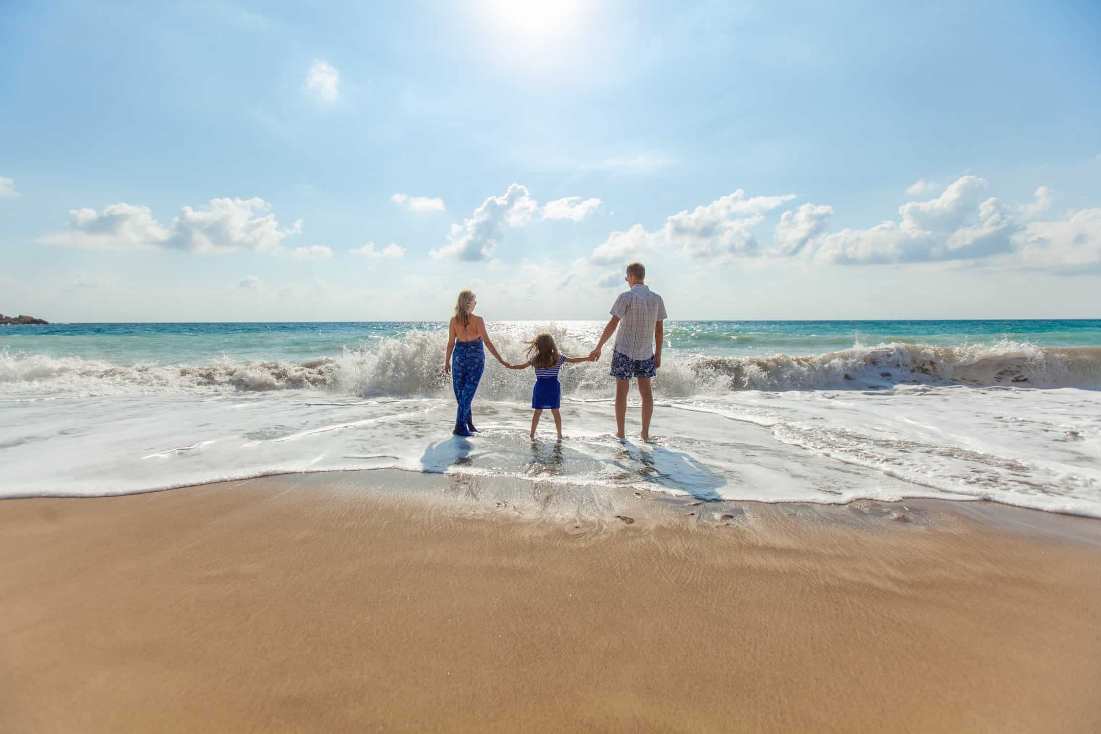 Man and woman holding hands of little girl whilst paddling in the sea with blue skies and golden sandy beach